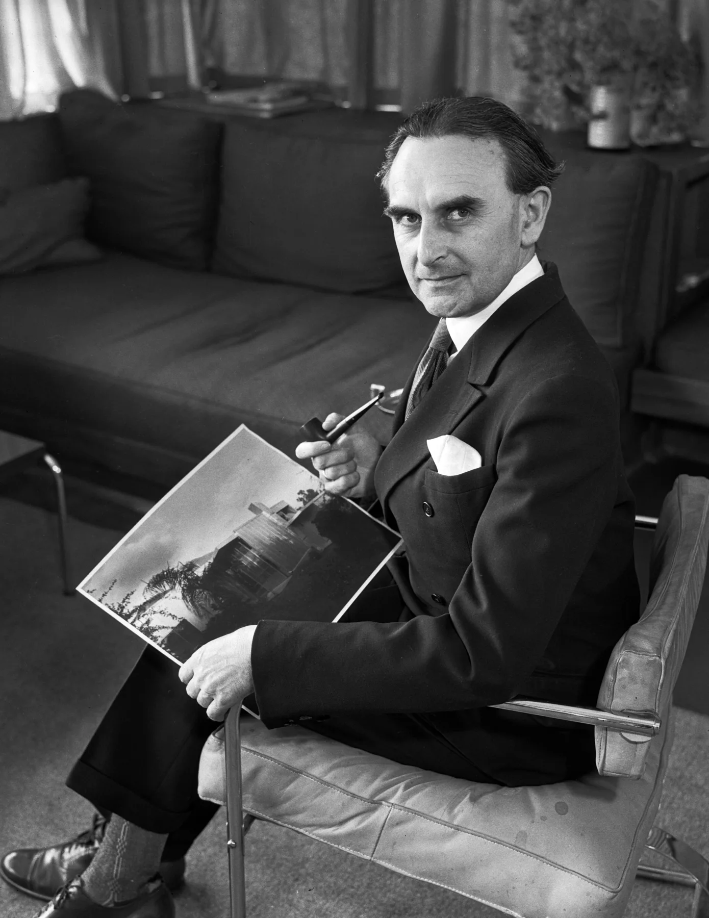 Richard Neutra seated in a tubular steel chair, holding a photograph of the Beard House, 1935.
