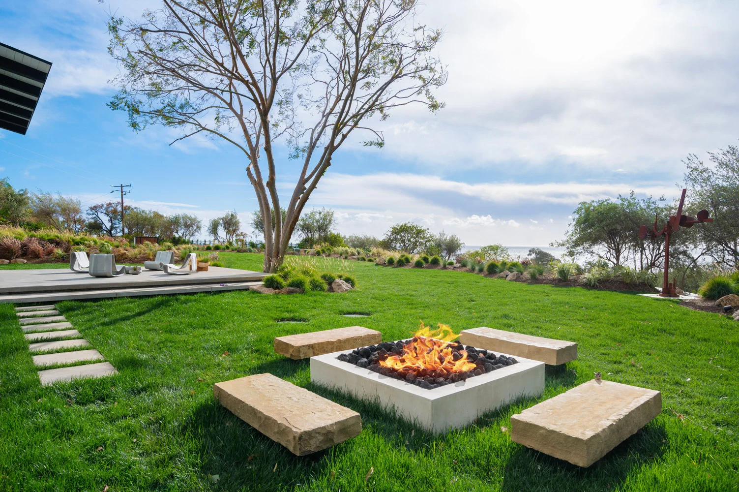The fire pit in the lawn, lit, under a spreading tree.