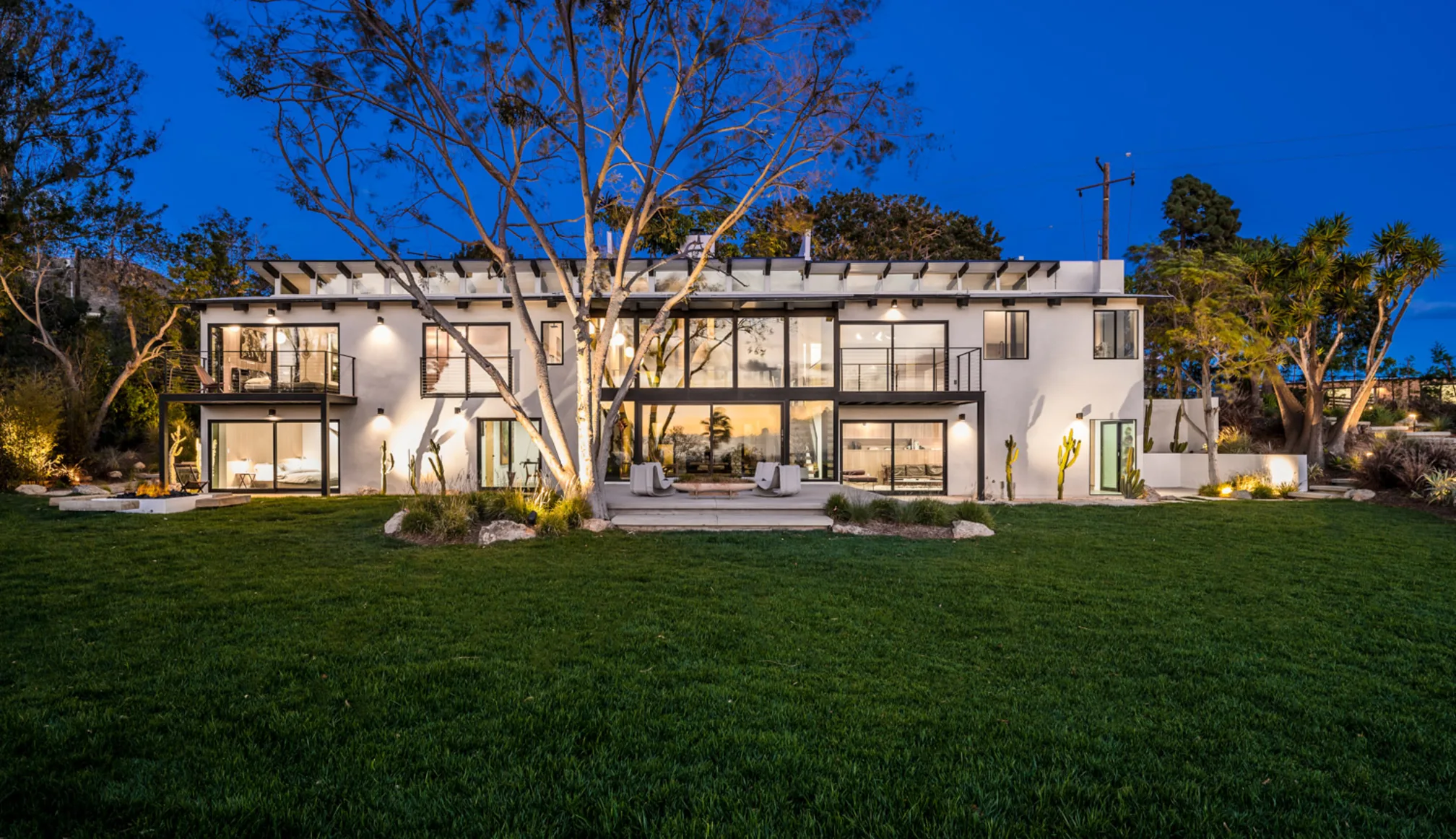 Filaree Heights, Malibu: the two-storey glass house lit from within at dusk, across its lawn.